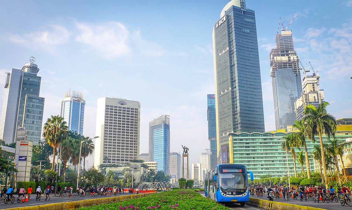 Cityscape with modern buildings, a blue bus, cyclists on the street, and clear skies.