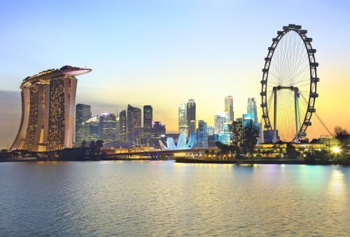 The image shows a stunning evening view of the Singapore skyline, featuring iconic landmarks like the Marina Bay Sands hotel and the Singapore Flyer ferris wheel, beautifully illuminated against a serene waterfront.