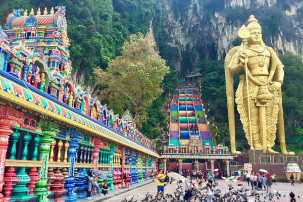 A golden statue of Lord Murgar stands majestically in Batu Caves, surrounded by lush greenery and vibrant temple architecture.