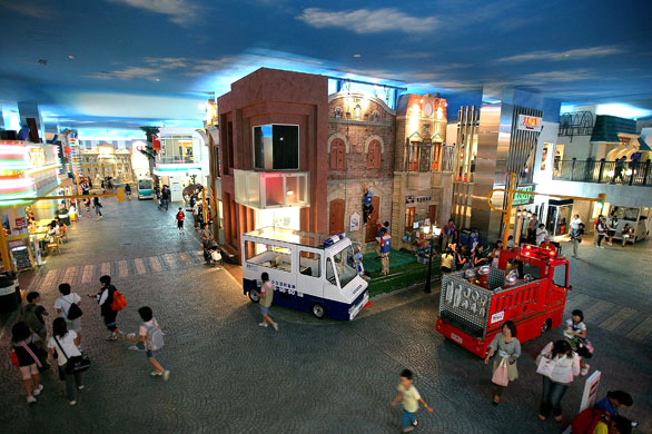 A lively indoor activity center designed to look like a city, with realistic buildings, a toy-like police car, and a fire truck. Visitors, including several families, walk through the streets, enjoying the interactive setup beneath a ceiling painted like the sky.