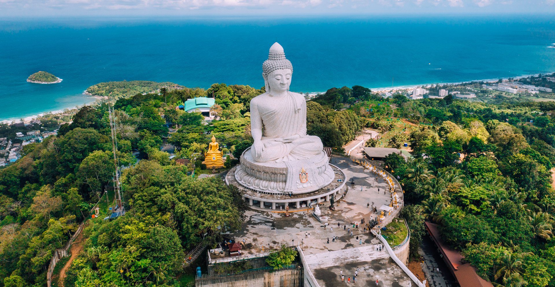 Aerial view of a large white seated Buddha statue surrounded by lush greenery and with a coastline in the background.