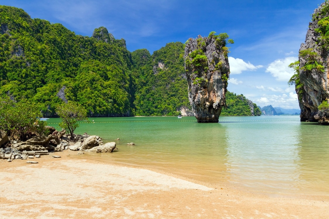 Tropical beach scene with clear water, sandy shore, large rock formations, and lush green cliffs under a blue sky.