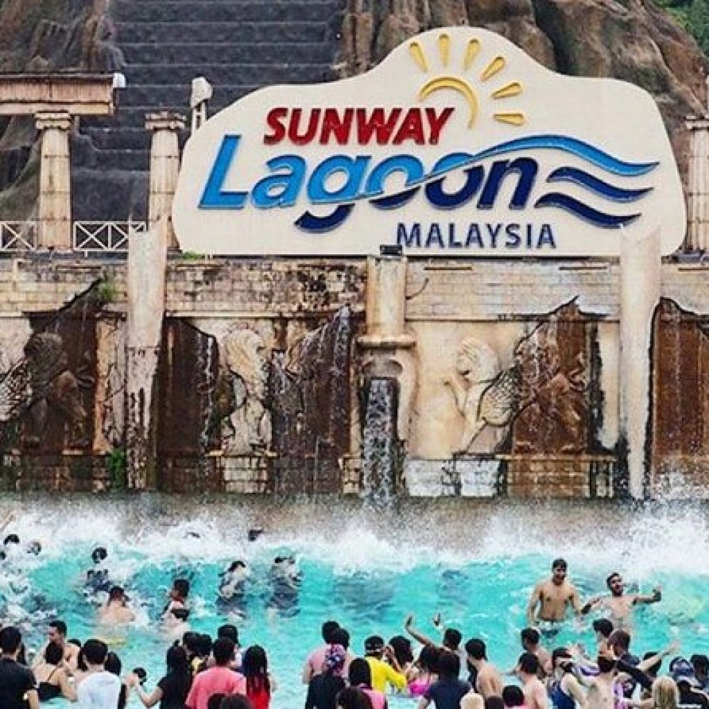 A lively scene at Sunway Lagoon in Malaysia, with a large crowd of people enjoying the wave pool in front of a grand stone structure. The Sunway Lagoon logo is prominently displayed above, with waves crashing behind the swimmers.