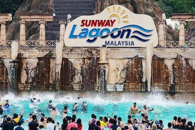 A lively scene at Sunway Lagoon in Malaysia, with a large crowd of people enjoying the wave pool in front of a grand stone structure. The Sunway Lagoon logo is prominently displayed above, with waves crashing behind the swimmers.