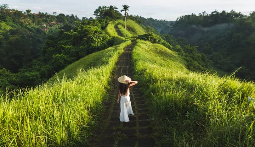 A woman in a white dress walks along a road surrounded by lush jungle foliage.