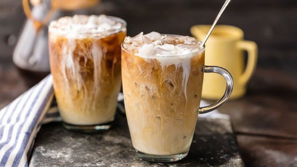 Two glasses of iced coffee with cream on a wooden surface next to a yellow mug.