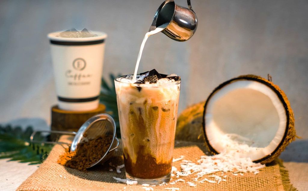 Iced coffee being poured into a glass with ice cubes, next to an open coconut and a cup on burlap.