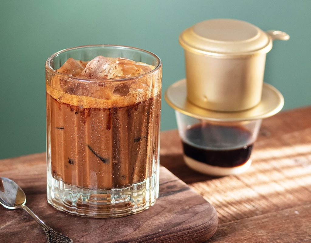 A glass of iced coffee with a Vietnamese drip filter beside it on a wooden surface.