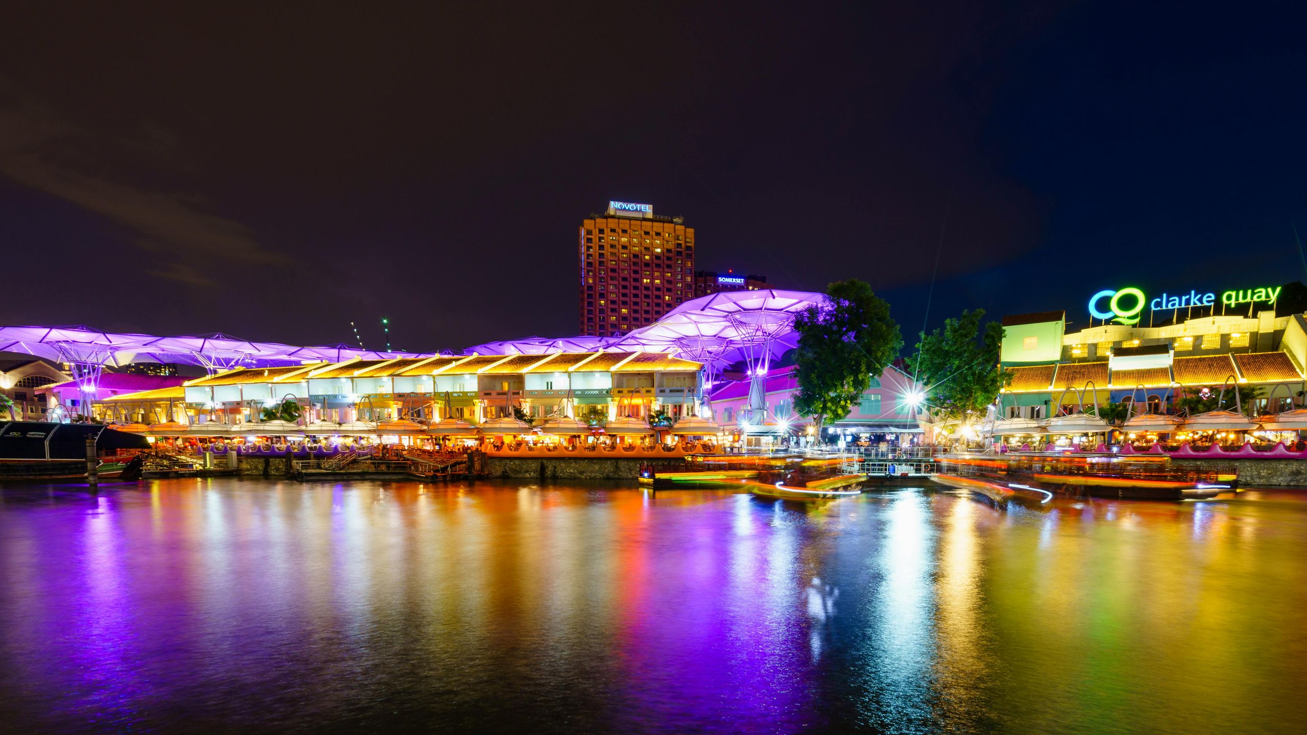 A serene body of water at night, reflecting the colorful lights of buildings along its banks in Clarke Quay. The scene captures the shimmering skyline and vibrant atmosphere, with a mix of architecture illuminated against the dark sky.