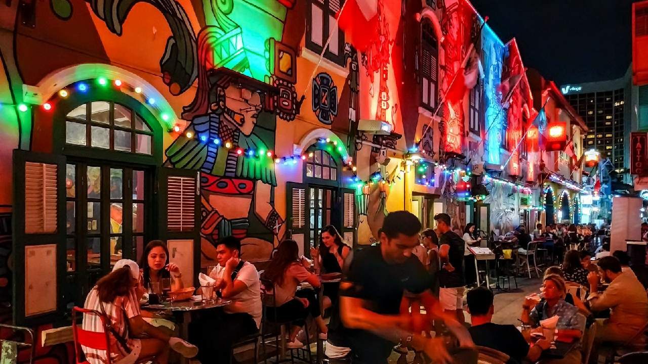 A group of people sitting at tables in an outdoor restaurant at night, with street lights illuminating the crowded scene in a city setting.