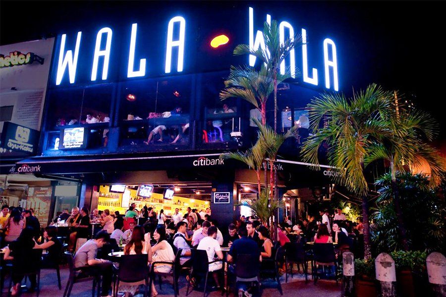 A group of people sitting outside a building at night, with neon signs for "WALT," "MALA," and "citibank" visible. The setting appears to be a food court near a casino.