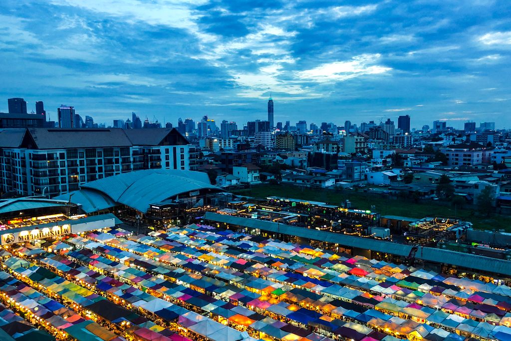 Aerial view of a vibrant night market with colorful tents, set against a city skyline during dusk.