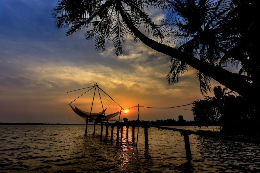 A boat is moored at a dock, surrounded by calm water under a clear sky with scattered clouds. In the background, trees line the shore of the lake, while the sun sets on the horizon, casting warm hues over the landscape in an evening scene.