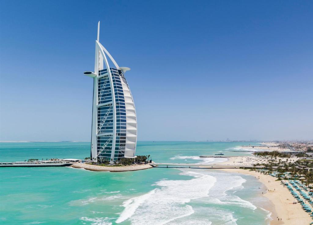 A sail-shaped building, the Burj Al Arab, stands on an island surrounded by turquoise sea and white sand beach under a clear blue sky.