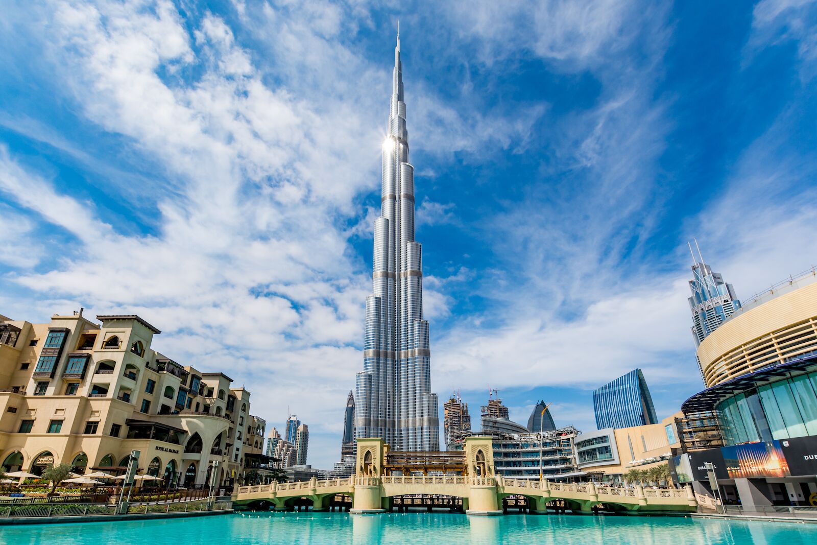 Tall skyscraper with a spire reaching toward the sky, surrounded by modern buildings and a pool in the foreground under a partly cloudy sky.