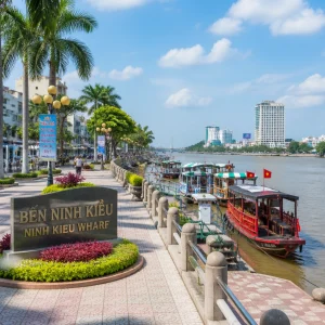 Ninh Kieu Wharf riverside promenade, boats along Hau River, palm-lined walkway, modern city surroundings, bright daylight, lively yet clean waterfront view